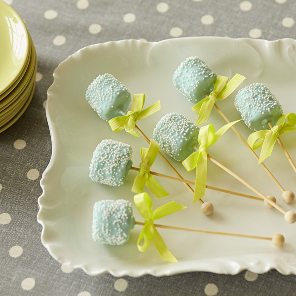 Blue marshmallow rattles on a stick decorated with blue candy melts, white sprinkles and green bows.