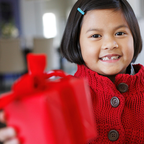 A little girl in a bright red coat is reaching out toward the camera with a wrapped gift in hand.