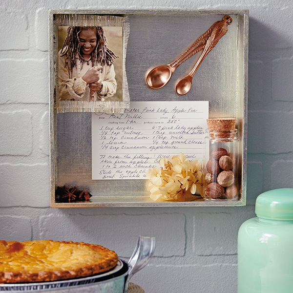 A shadow box containing a recipe card, a photo of a woman smiling, a small jar of herbs, and two decorative measuring spoons hangs on a white brick wall near a baked pie in a pie tin and a vase of white flowers.