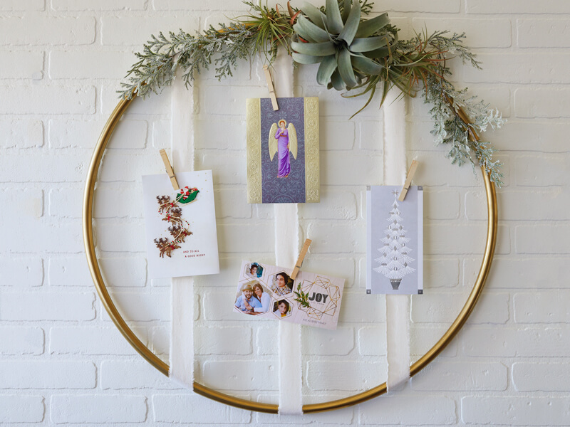 Gold hoop Christmas card holder with greenery on the outside and cards held into place by vertical ribbons and clips.