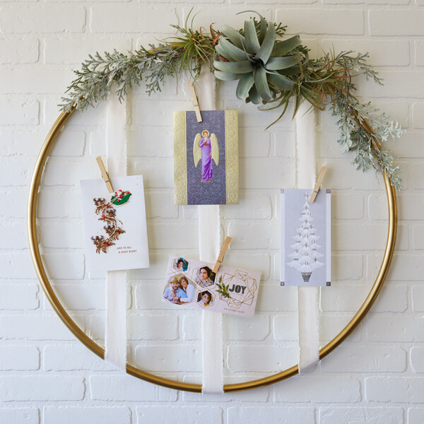A gold hoop Christmas card display with greenery on the top and vertical white ribbons.