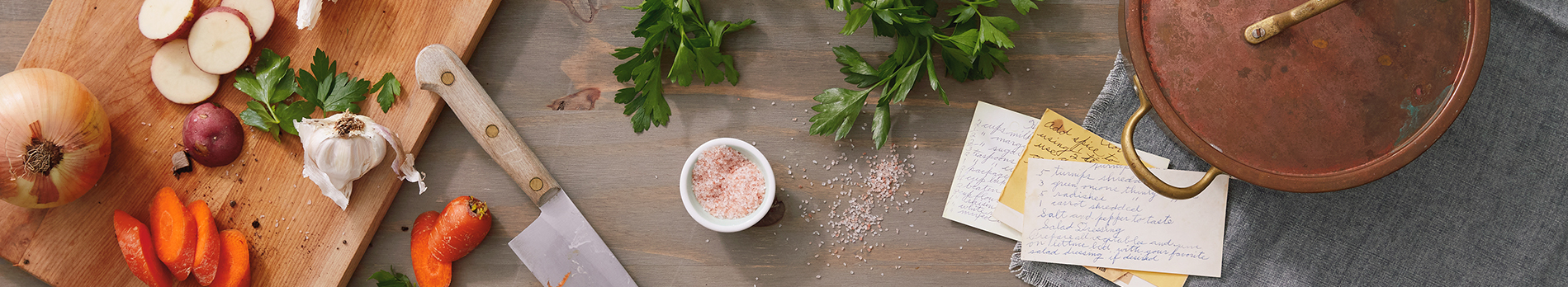 Old recipe cards with handwritten recipes fanned out on a kitchen counter with herbs and a copper pot.