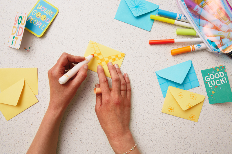 Hands drawing patterns on a Just Because Minis envelope with Crayola markers on a speckled countertop.