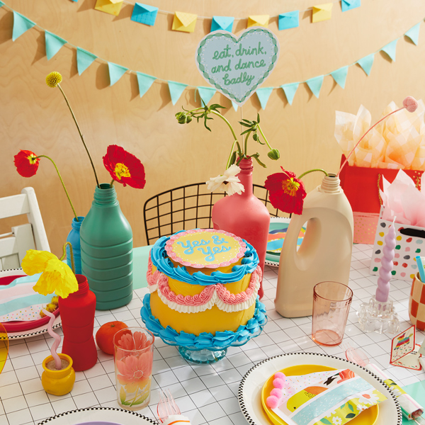 A table set with DIY party decorations, including a cake with a free printable cake topper, a mini card envelope garland, and cake favor pouches.