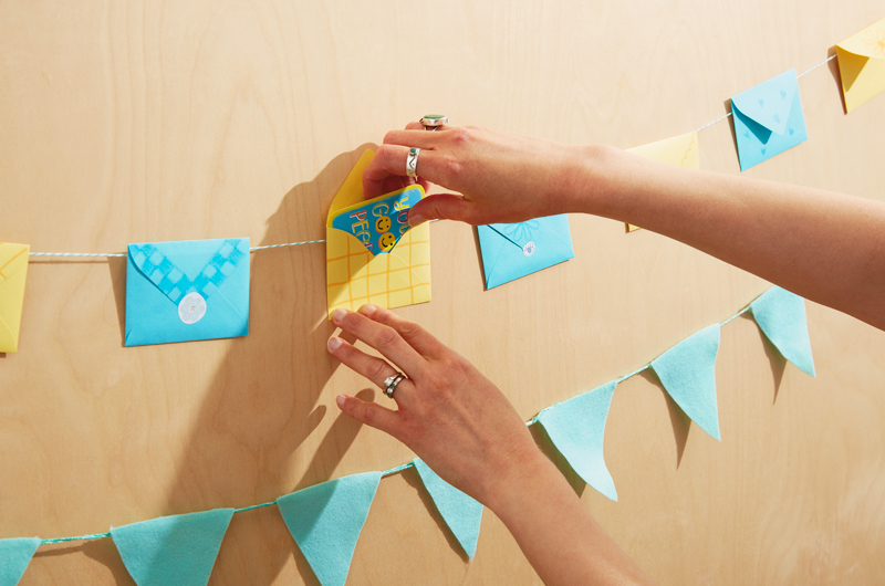 Hands reaching into frame to place a Just Because Mini card in an envelope that's attached to a garland strung with multiple mini envelopes.