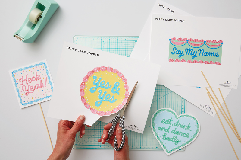 Hands cutting out a free printable cake topper that reads,