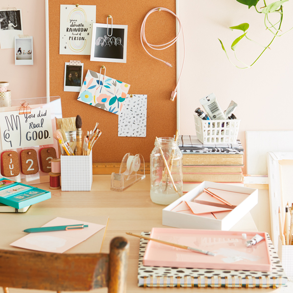 A writing desk covered with journals, pens, markers and paper; on the wall in front of the desk is a bulletin board covered with notes and pictures.