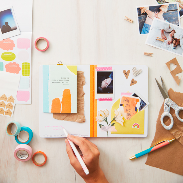 A hand writing in a mixed media journal, surrounded by art supplies like Washi tape, scissors and markers.