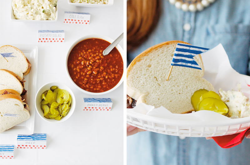 Fourth of July food labels and flags on a table and sticking out of a sandwich.