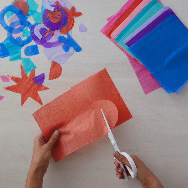 Hands cutting shapes out of brightly colored sheets of tissue paper with a pair of scissors.