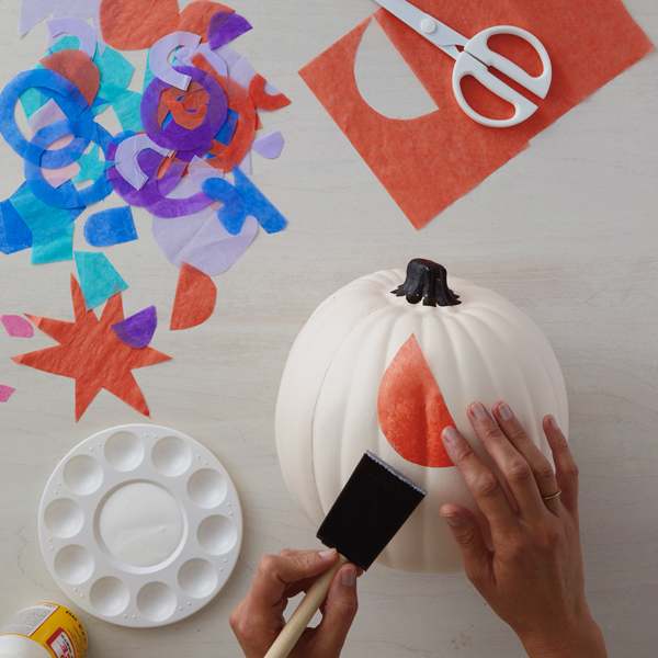 Hands decoupaging an orange half-moon shape tissue paper cut out onto a white pumpkin with a foam brush.