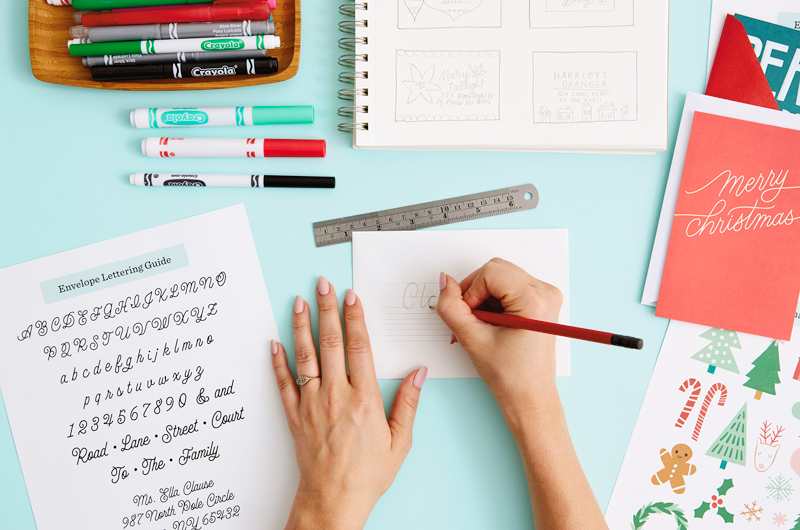 A woman's hands demonstrating how to use the Envelope Lettering Guide to letter an envelope, penciling letters onto lightly sketched ruled lines.