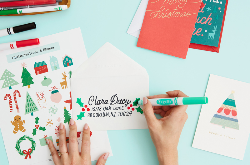 A woman drawing little holly branches on her hand lettered envelope.