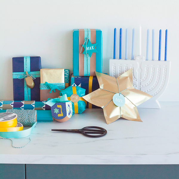A pile of Hanukkah gifts on a table, wrapped in paper in varying shades of blue and gold, with a white ceramic menorah with blue candles in the background.