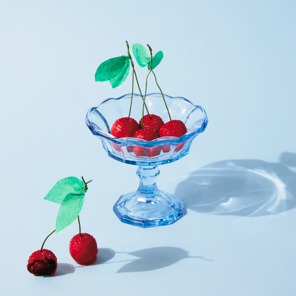 Chocolate cherry cake truffles decorated to look like real cherries sit in a clear blue glass dessert dish with a scalloped rim.