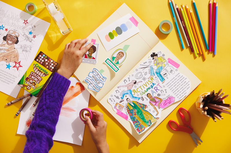 A scrapbook lays on a bright yellow surface. A child's hands are reaching into frame, taping portions of the Black History Month coloring pages inside the scrapbook. Scattered around the work surface are various coloring tools, tape and scissors.