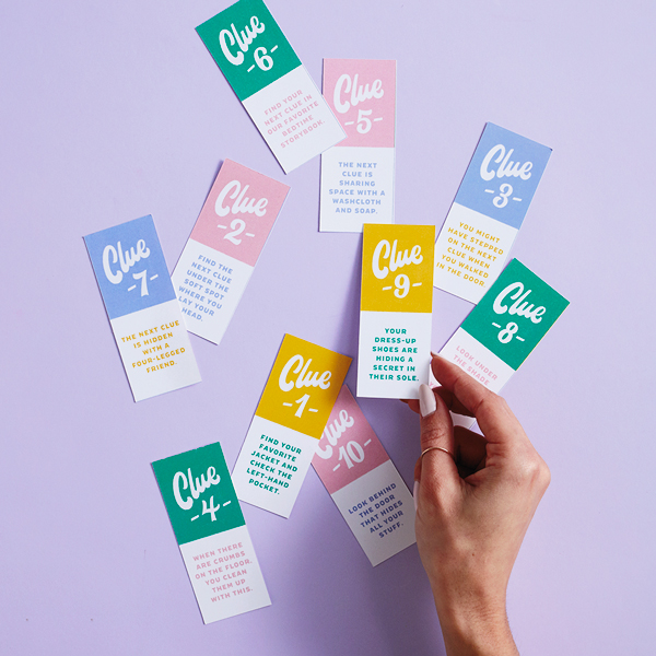 A hand reaches in to pick up one of the scavenger hunt clue slips from amid a bunch of clue slips scattered on a light purple surface.