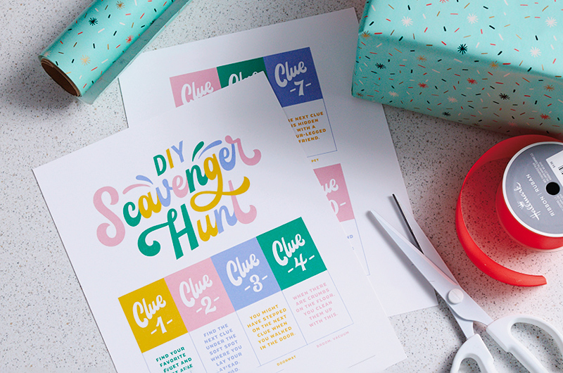 The scavenger hunt free printable sits on a desktop next to a wrapped gift, some ribbon and a pair of scissors.