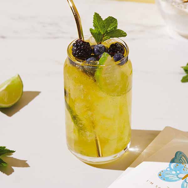 A glass of green tea soda garnished with blackberries, lime and mint, with a gold-plated metal straw, sitting on a white and gray marble countertop with a Signature butterfly card sitting nearby.