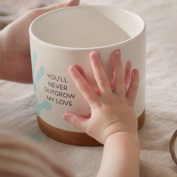 A toddler places its hand on a pink handprint on the side of a planter handprint kit gift for Mother's Day.
