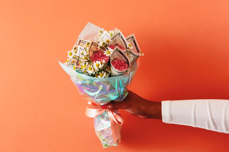 A woman's hand reaches into frame with a bouquet of flowers; multiple flower heads are wrapped in five and ten dollar bills.