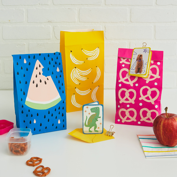Three paper lunch sacks—a blue one, a yellow one, and a bright pink one—are decorated with cut paper shapes of different snacks, including watermelon, bananas and pretzels; they're sitting on a white countertop in front of a white brick background, with a container of mini pretzels, an apple, and a Just Because mini card sitting nearby.
