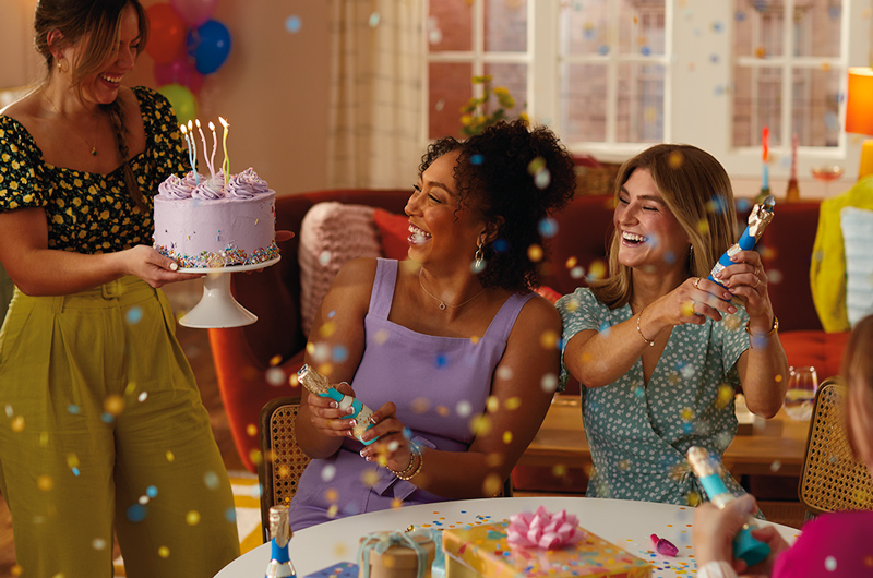 A woman in a purple dress sits in the middle of two friends at a birthday party; the friend on her right is holding out a purple iced birthday cake with lit candles on a cake stand toward her, while the friend on her left is holding a confetti popper up in the air, having just popped it. Confetti is raining down in the foreground.