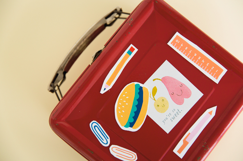 Our free, DIY back-to-school magnets clinging to the side of a red metal lunchbox, holding a note that features an illustration of two pieces of fruit and the message,