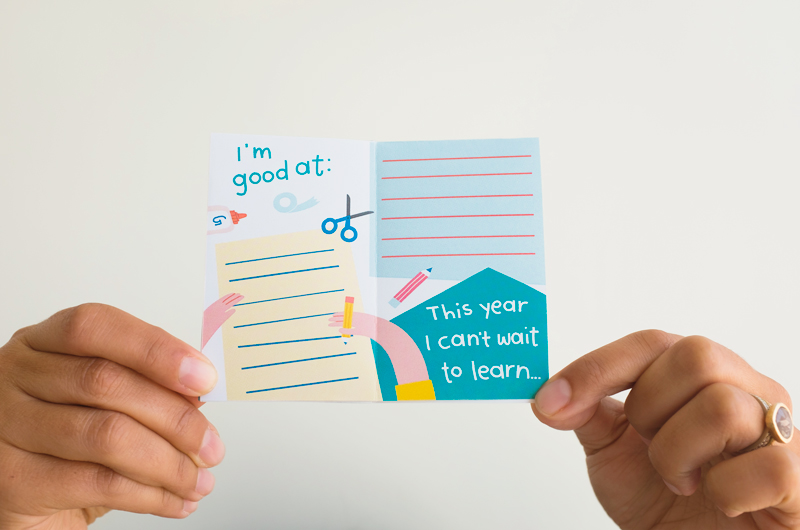 A woman's hands hold up the inside of an assembled back-to-school zine with fill-in-the-blank spaces for kids that read,