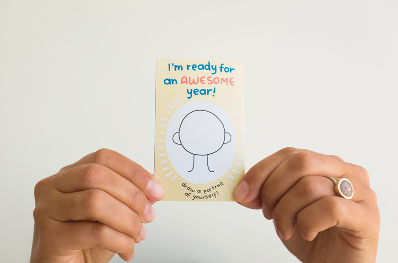 A woman's hands hold up the back of a back-to-school zine with a blank face for a child to draw their portrait on and the headline,