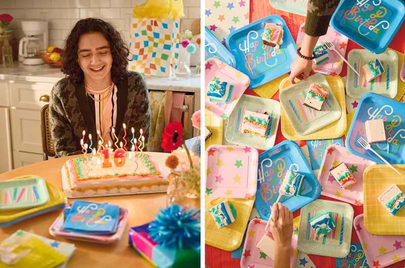 A teenage boy sits at a kitchen table, looking down and smiling at an iced birthday cake with a lit 