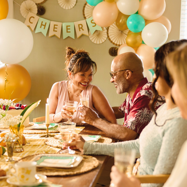 A husband and wife celebrating their wedding anniversary sit at the end of a party table, clinking their vintage glasses of champagne in a toast; the table is set with Celebrate! partyware and behind them hangs a party banner in shades of pink, light aqua and gold that reads,