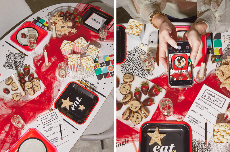 A downshot of an awards night party table, set with red, white and black Celebrate! partyware and scattered with snacks like popcorn, puffed rice cereal treats, chocolate-covered strawberries, cookies, and personal awards show voting ballots; next to this, a different angle of the scene is being captured by a pair of hands snapping a photo with a smartphone.