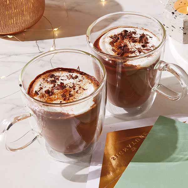 Two clear mugs filled with honey hot chocolate and garnished with chocolate shavings sit on a white and gray marble countertop; fairy lights and a Signature card envelope lay nearby.