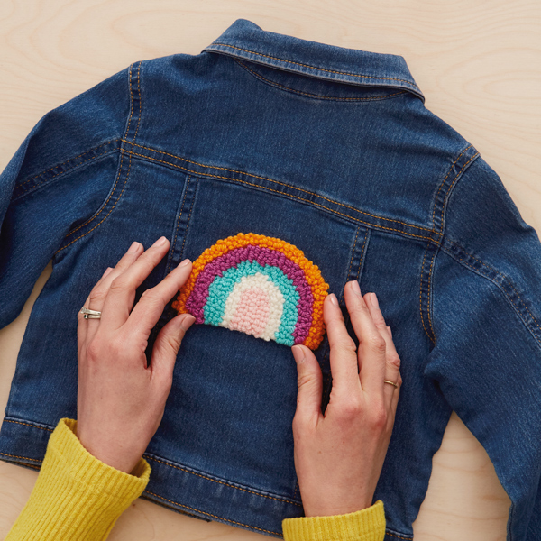 A woman's hands center a punch needle embroidery patch in the shape of a brightly colored rainbow onto the back of a dark blue denim jacket.