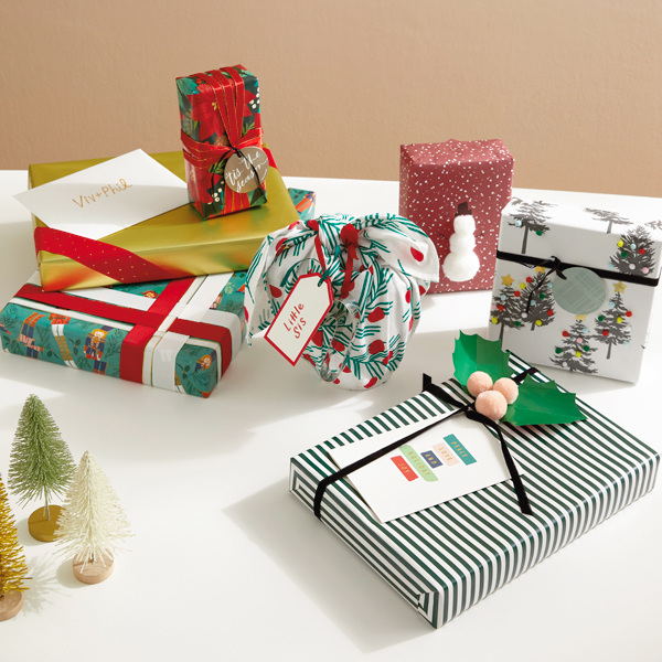 Three different Christmas gift wrapping ideas are shown via differently wrapped gifts sitting on a white tabletop; one gift is wrapped and embellished with woven ribbons, another is wrapped and decorated with a simple ribbon DIY made to look like a holly sprig, and a final gift is wrapped with fabric covered in a simple holly print.