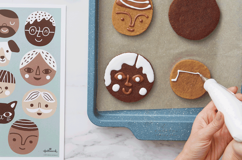 A stop-motion GIF of a woman using a piping bag full of white royal icing to draw a face and hair on a round gingerbread cookie; next to the cookie sheet on the white marble countertop lays a printout of a guide for drawing different facial features.