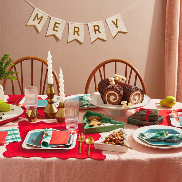 A Christmas tablescape featuring Hallmark Celebrate! partyware products like dark green hexagon-shaped plates and dark green and white plaid napkins; at the center of the table is a traditional yule log cake covered in chocolate frosting and meringue marshmallows, sitting on a simple white cake stand; nearby is a set of mod gold candle holders with white, twisted candles in them; behind the scene is a banner that says
