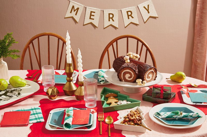 A Christmas tablescape featuring Hallmark Celebrate! partyware products like dark green hexagon-shaped plates and dark green and white plaid napkins; at the center of the table is a traditional yule log cake covered in chocolate frosting and meringue marshmallows, sitting on a simple white cake stand; nearby is a set of mod gold candle holders with white, twisted candles in them; behind the scene is a banner that says