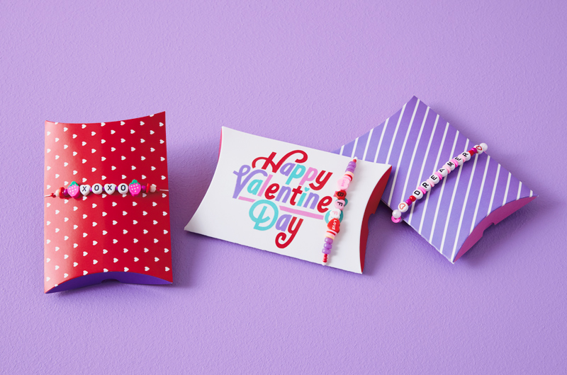 Three paper pillow boxes are decorated with handmade friendship bracelets that have been stretched around them; the pillow boxes feature different patterns like red with tiny white hearts, lavender with white diagonal stripes, and a white box with