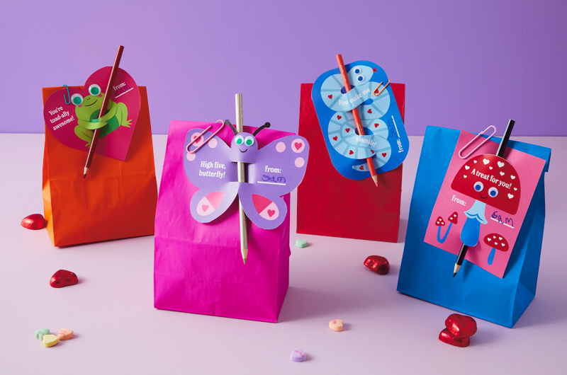 Assembled Valentine's Day treat bags featuring our free printable Valentine's Day friends sit assembled on a light purple surface scattered with heart-shaped chocolates and candy conversation hearts; the free printables include a snake, a toadstool mushroom, a butterfly and a toad, all with a colored pencil stuck through them to
