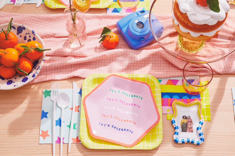 A party table set with plates and napkins in cheerful, bright shades of orange, yellow, pink, blue and green; next to the plate is a DIY personalized photo place holder that's been painted in bright colors and has a small Instax photo pasted inside the frame; a blue Instax camera sits at the center of the table between a bowl of mandarin oranges and a transparent yellow glass cake stand with a cake piled high with meringue frosting and a mandarin orange on top.