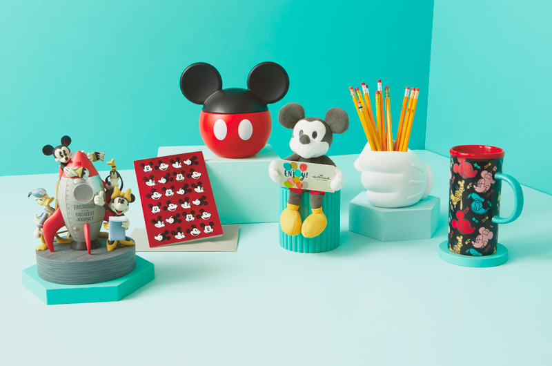A selection of gifts meant for a parent who loves Disney, including a small plush Disney Mickey Mouse gift card holder, a sculpted pencil holder in the shape of Mickey's gloved hand, a color-changing travel mug featuring the faces of Mickey Mouse, Minnie Mouse, Donald Duck and Goofy; a treat jar with a lid that looks like black Mickey Mouse ears; a figurine of Mickey, Minnie, Donald and Goofy prepping a vintage-looking spaceship for launch; and a greeting card featuring the face of Mickey Mouse in a variety of different expressions on top of a red background.