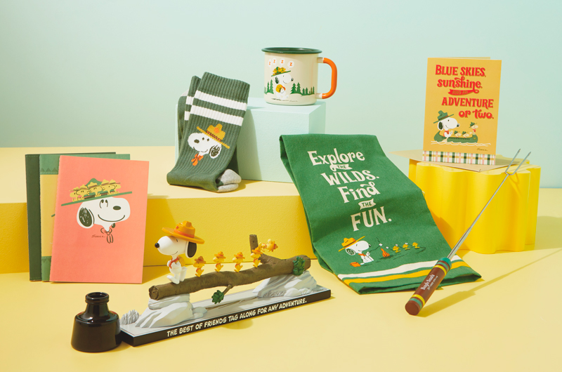 A selection of gifts for a parent who loves Peanuts Snoopy, specifically the Beagle Scouts, including a set of three soft-cover Beagle Scout journals, a desktop figurine that features Snoopy in his Beagle Scout uniform leading a troop of yellow canaries across a fallen log, Woodstock trailing in the background; the figurine reads