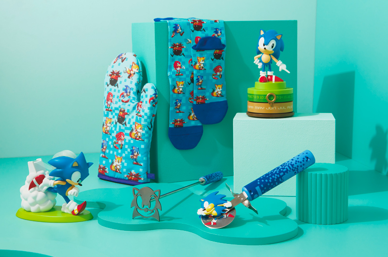 A selection of gifts for parents who love Sonic the Hedgehog that includes a Sonic the Hedgehog perpetual calendar, a rotary pizza cutter, a grill brand in the shape of Sonic the Hedgehog's face; a tape dispenser featuring a figurine of Sonic running and leaving a cloud of dust in his wake; a pair of blue crew socks and a blue oven mitt covered in 8-bit style illustrations of Sonic, Knuckles, Miles (aka