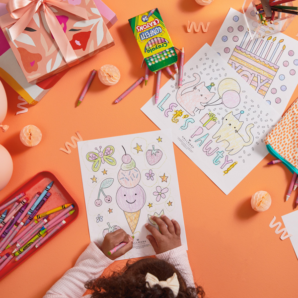 A little girl with a white bow in her hair colors in a free printable birthday coloring page on a coral-colored surface; surrounding her are more coloring pages, a tray full of crayons in different hues, gift boxes in shades of orange, white, pink and red, and light pink balloons.