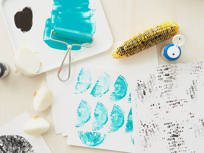 A cob of corn and quarters of an onion have been coated with paint and rolled across pieces of paper to create interesting textures and shapes; the onion was painted with turquoise paint and has left half-moon shapes with arcs inside, like a rainbow, while the corn has been coated in red and black paint and left a pattern that looks like closely spaced polka dots.