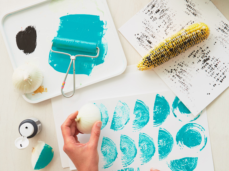 cob of corn and quarters of an onion have been coated with paint and rolled across pieces of paper to create interesting textures and shapes; the onion was painted with turquoise paint and has left half-moon shapes with arcs inside, like a rainbow, while the corn has been coated in red and black paint and left a pattern that looks like closely spaced polka dots.