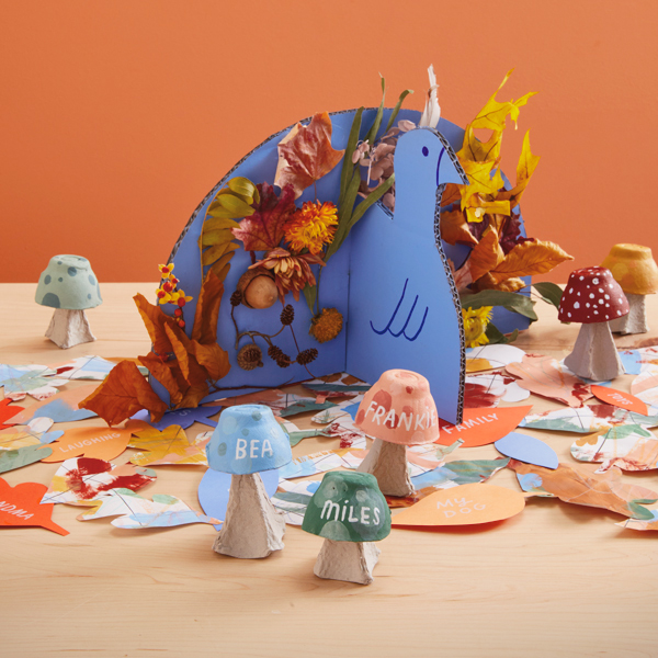 A DIY Thanksgiving centerpiece in the shape of a turkey and decorated with foraged fall objects like fallen leaves and acorns sits on a table top; surrounding it are additional kid-friendly Thanksgiving crafts, such as egg carton mushroom placeholders and a gratitude table runner made from cut-out paper leaves.