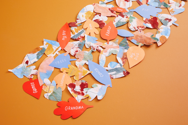 A gratitude table runner made with leaf-shaped cut-outs of wrapping paper and construction paper; written on the solid-colored pieces of construction paper are words like,